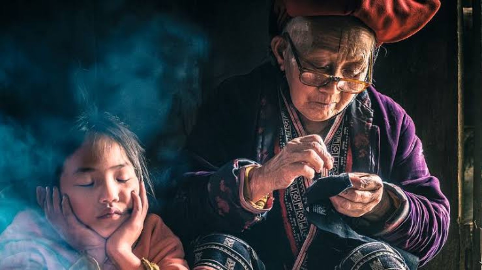An older woman sewing by hand beside a young child, in soft window light.